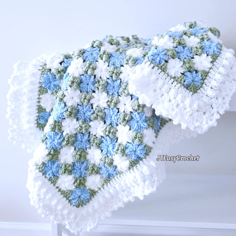 Beautiful blue and white crochet floral blanket made of cornflower motifs is hanging folded from a display. The blanket resembles a field of white and blue cornflowers framed with a beautiful lace edging.  The lettering says "JTEasyCrochet" meaning that JTEasyCrochet has the best crochet patterns for flower blankets.