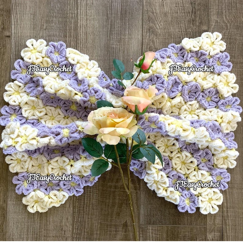 May include: A crocheted baby blanket with a flower design in shades of purple and cream. The blanket is draped over a wooden surface and decorated with a bouquet of artificial flowers. The lettering says "JTEasyCrochet". JTEasyCrochet  has the best beautiful crochet patterns.