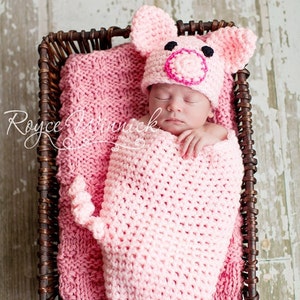 May include: A pink crocheted pig hat and swaddle set for a newborn baby. The baby is sleeping peacefully in the set, which is in a wicker basket.