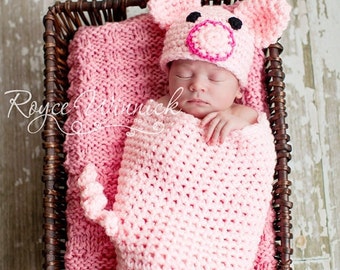 May include: A pink crocheted pig hat and swaddle set for a newborn baby. The baby is sleeping peacefully in the set, which is in a wicker basket.