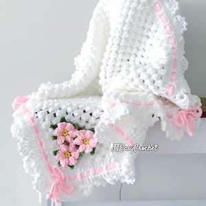 May include: A white crocheted blanket with a textured pattern and pink detailing. The blanket has a floral design with pink flowers and green leaves. The edges are trimmed with a pink ribbon and a scalloped border. The text "JTEasyCrochet" is visible.