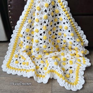 Op de afbeelding: Een witte gehaakte deken met een madeliefpatroon, met gele centra en een gele rand. De deken is gemaakt van zacht garen en heeft een geschulpte rand. De tekst "JTEasyCrochet" is zichtbaar in de linker benedenhoek.