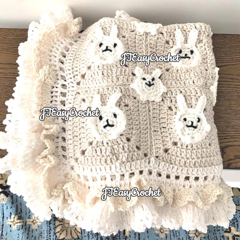 Op de afbeelding: Een gehaakt babydekentje met een cr&egrave;mekleurige achtergrond en witte konijnenapplicaties. Het dekentje heeft een gerimpelde rand en de tekst "JJ Easy Crochet" is zichtbaar op de afbeelding.