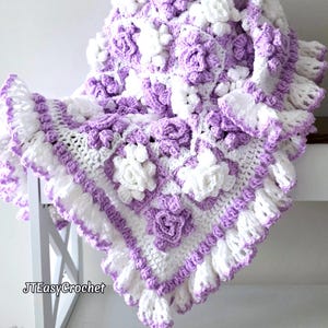 May include: A handmade crochet blanket in white and lavender, featuring a floral pattern with raised rose appliqués. The blanket has a ruffled edge and the text "JTEasyCrochet" is visible.