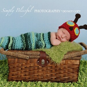 May include: A newborn baby wearing a red, green, and yellow crocheted caterpillar hat and a green and blue striped crocheted sleeping bag, lying on a green blanket in a brown wicker basket.