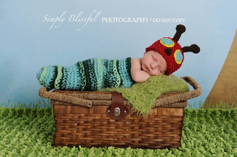 May include: A newborn baby wearing a red, green, and yellow crocheted caterpillar hat and a green and blue striped crocheted sleeping bag, lying on a green blanket in a brown wicker basket.