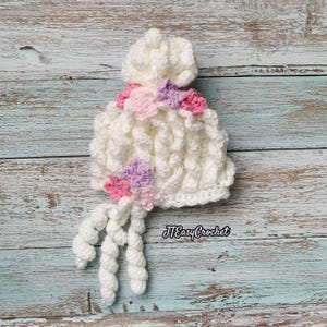 May include: A white crochet hat with a textured pattern, adorned with pink and purple crochet flowers. The hat has a unique design with dangling, twisted yarn accents. The text "JTEasyCrochet" is visible on the bottom right.