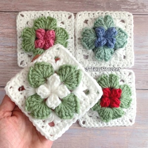 May include: Four crocheted squares featuring different coloured flowers in the centre. The flowers are made with green and white yarn, and the centres are pink, blue, red, and white. The squares are all white and have a delicate crocheted pattern.