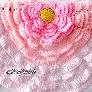 May include: A handmade crochet flower in shades of pink, with a yellow centre. The flower is the focal point of a larger, scalloped design, also in varying shades of pink. The text "JTEasyCrochet" is visible in the lower left corner.