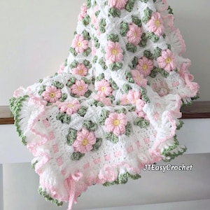 May include: A crocheted blanket with a floral pattern. The blanket is white with pink and yellow flowers, green leaves, and a ruffled edge. The text "JTEasyCrochet" is visible on the blanket.