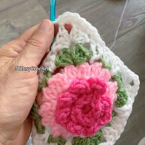 A hand is holding a crochet hook and a white granny square close up with shades of pink peony in the center of the square with green leaves in the corners.