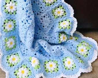 May include: A handmade, light blue crochet blanket featuring daisy motifs. Each daisy has white petals, a yellow center, and green leaves. The blanket has a scalloped white border and the text "JTEasyCrochet".