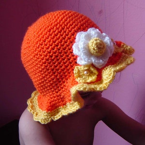 A crocheted hat in orange with a yellow and white flower design. The hat has a scalloped brim.