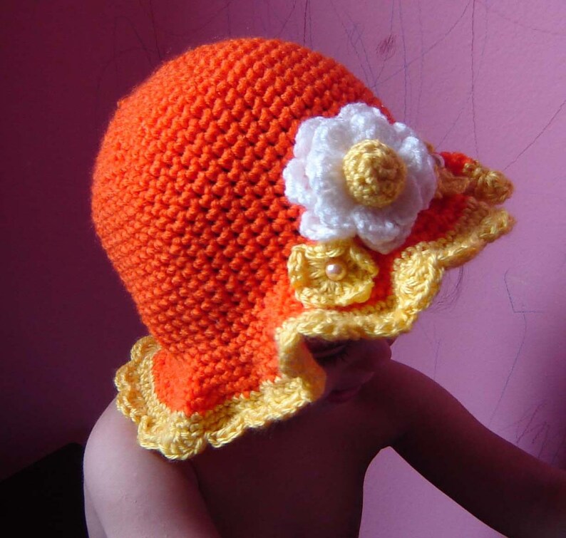 A crocheted hat in orange with a yellow and white flower design. The hat has a scalloped brim.