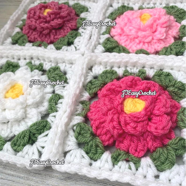 Clode up on set of four piled up 3D white granny squares with raised borders laying on a flat surface. Each granny square has a white or shades of pink rose or peony with 2 layers of petals surrounded with green leaves.