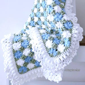 Lavishly beautiful crochet floral blanket made of cornflower motifs is hanging from a display. The blanket resembles a field of white and blue cornflowers framed with a beautiful lace edging.  The lettering says "JTEasyCrochet" meaning that JTEasyCrochet has the best crochet patterns for flower blankets.