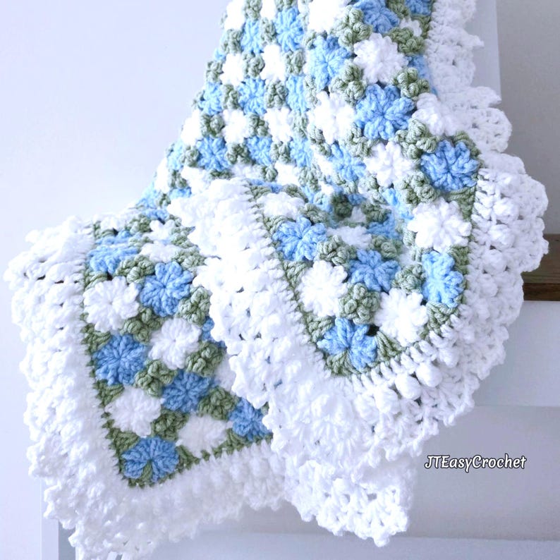 Lavishly beautiful crochet floral blanket made of cornflower motifs is hanging from a display. The blanket resembles a field of white and blue cornflowers framed with a beautiful lace edging.  The lettering says "JTEasyCrochet" meaning that JTEasyCrochet has the best crochet patterns for flower blankets.