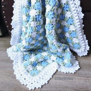 May include: A handmade crochet blanket in shades of white, light blue, and green. The blanket features a floral pattern and a scalloped white border. The text "JTEasyCrochet" is visible in the corner.