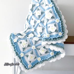 Beautiful crochet granny square blanket is hanging from a display on a light grey background. Each white granny square has a white textured gardenia flower in the center, framed with pale green leaves and light frosty blue border, so the whole blanket remind of a frozen gardenia blanket. There are also bunches of gardenia flowers in the image to highlight the likeness. The lettering logo says: " JTEasyCrochet"