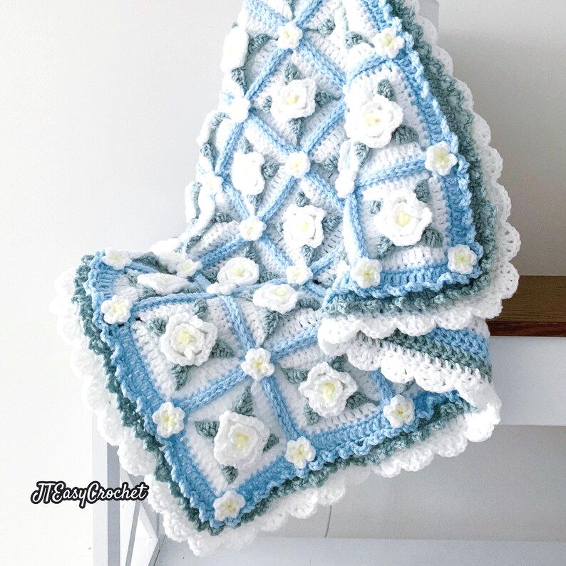 Beautiful crochet granny square blanket is hanging from a display on a light grey background. Each white granny square has a white textured gardenia flower in the center, framed with pale green leaves and light frosty blue border, so the whole blanket remind of a frozen gardenia blanket. There are also bunches of gardenia flowers in the image to highlight the likeness. The lettering logo says: " JTEasyCrochet"