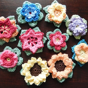 May include: A collection of ten crocheted flowers in various colours, including pink, red, blue, yellow, and purple. The flowers are all different sizes and shapes, and they are arranged in a circle on a wooden surface.