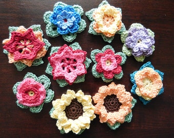 May include: A collection of ten crocheted flowers in various colors, including pink, red, blue, yellow, and purple. The flowers are all different sizes and shapes, and they are arranged in a circle on a wooden surface.