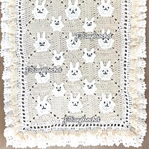 Op de afbeelding: Een gehaakt babydekentje met een beige achtergrond en witte konijnenapplicaties. Het dekentje heeft een witte gehaakte rand met een gekrulde zoom.