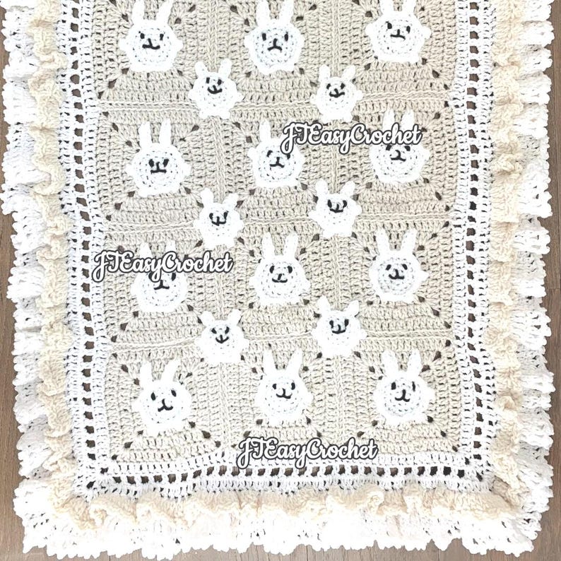 Op de afbeelding: Een gehaakt babydekentje met een beige achtergrond en witte konijnenapplicaties. Het dekentje heeft een witte gehaakte rand met een gekrulde zoom.