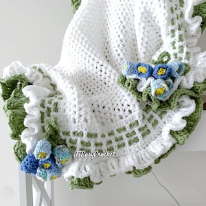 Pode incluir: Um cobertor de bebé de crochet branco com uma borda de crochet verde e flores de crochet azul e branco. O cobertor tem uma borda com babados. O texto "JJ Easy Crochet" é visível no canto inferior direito da imagem.