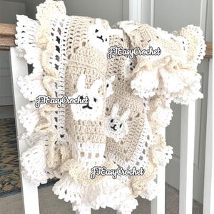 Op de afbeelding: Een gehaakt babydekentje in wit en beige met een gegolfde rand. Het dekentje heeft een herhalend patroon van konijntjes. Het dekentje is op meerdere plekken met "JJ Easy Crochet" gelabeld.