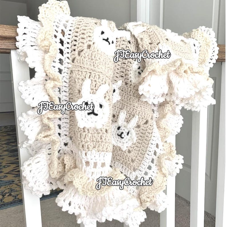 Op de afbeelding: Een gehaakt babydekentje in wit en beige met een gegolfde rand. Het dekentje heeft een herhalend patroon van konijntjes. Het dekentje is op meerdere plekken met "JJ Easy Crochet" gelabeld.