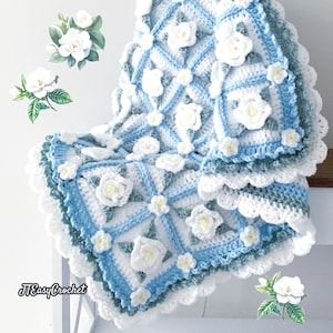 Beautiful crochet granny square blanket is hanging from a display on a light grey background. Each white granny square has a white textured gardenia flower in the center, framed with pale green leaves and light frosty blue border, so the whole blanket remind of a frozen gardenia blanket. There are also bunches of gardenia flowers in the image to highlight the likeness. The lettering logo says: " JTEasyCrochet"