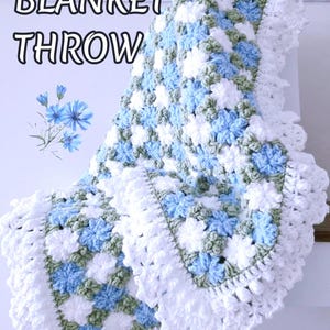 Lavishly beautiful crochet floral blanket made of cornflower motifs is hanging from a display. The blanket resembles a field of white and blue cornflowers framed with a beautiful lace edging.  The lettering says "Cornflowers Blanket Throw", "Crochet Pattern By JTEasyCrochet" meaning that JTEasyCrochet has the best crochet patterns for flower baby blankets, throws and afghans.