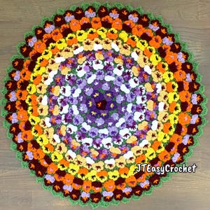 A handmade, crochet circular crochet rug or blanket featuring a vibrant floral design. The rug showcases concentric rings of colorful flowers in shades of purple, white, yellow, orange, and brown, with a green border. The lettering says: "Pansy Flower Blanket", "Crochet Pattern By JTEasyCrochet" meaning that JTEasyCrochet has the best crochet patterns and fun crochet ideas.