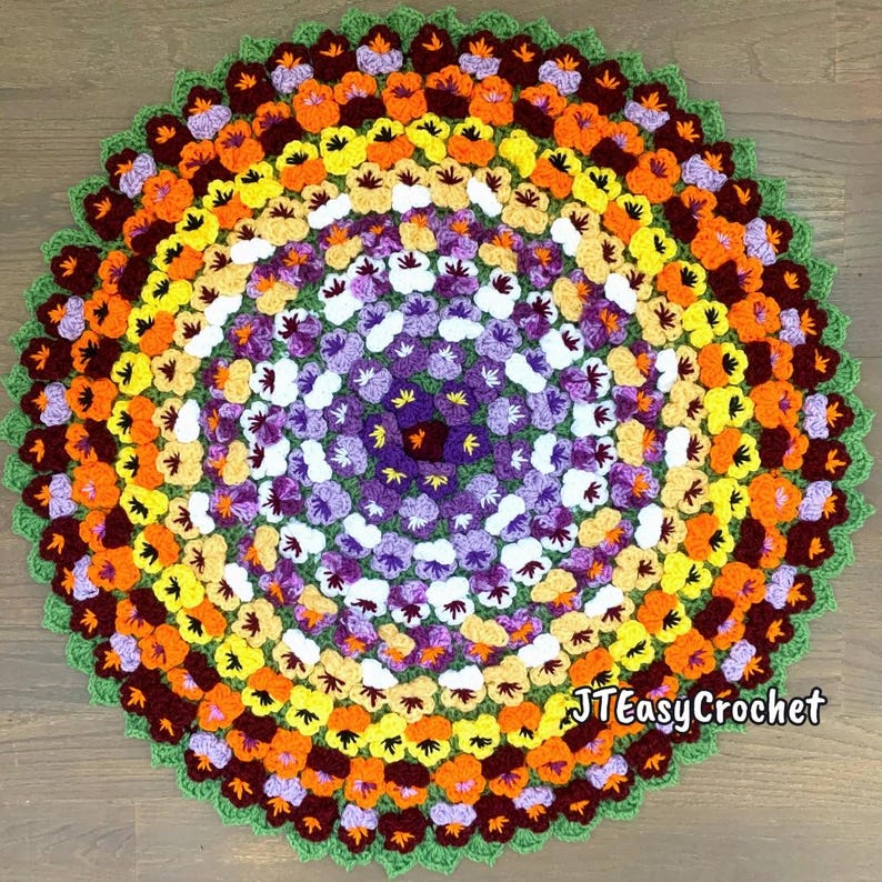 A handmade, crochet circular crochet rug or blanket featuring a vibrant floral design. The rug showcases concentric rings of colorful flowers in shades of purple, white, yellow, orange, and brown, with a green border. The lettering says: "Pansy Flower Blanket", "Crochet Pattern By JTEasyCrochet" meaning that JTEasyCrochet has the best crochet patterns and fun crochet ideas.