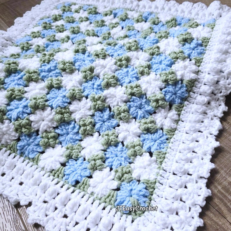 Beautiful blue and white crochet floral blanket made of cornflower motifs is laying on a wooden surface. The blanket resembles a field of white and blue cornflowers framed with a beautiful lace edging.  The lettering says "JTEasyCrochet" meaning that JTEasyCrochet has the best crochet patterns for flower blankets.