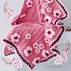 Könnte beinhalten: Eine handgefertigte rosa Häkeldecke mit 3D-Kirschblüten. Die Decke hat ein Rippenmuster und einen dunkelburgunderfarbenen und weißen gewellten Rand. Die Blüten sind in verschiedenen Größen und Rosatönen gehalten.