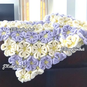 Puede incluir: Una manta de ganchillo hecha a mano con un patrón floral en tonos lavanda, crema y amarillo pálido. La manta está drapeada sobre una superficie oscura, mostrando el intrincado trabajo de puntadas y la paleta de colores. El texto "JJEasyCrochet" es visible.