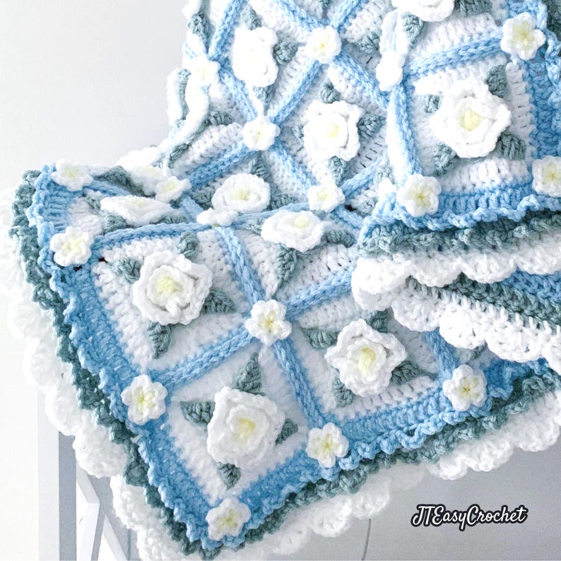 Beautiful crochet granny square blanket is hanging from a display on a light grey background. Each white granny square has a white textured gardenia flower in the center, framed with pale green leaves and light frosty blue border, so the whole blanket remind of a frozen gardenia blanket. There are also bunches of gardenia flowers in the image to highlight the likeness. The lettering logo says: " JTEasyCrochet"