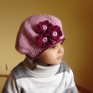 May include: A pink crocheted beret with a burgundy flower embellishment. The beret is worn by a child with a striped sweater.