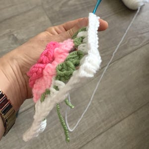 A hand is holding a floral granny square with 3D rose in the center with shades of pink petals and green leaves on a white square.