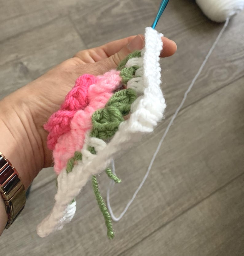 A hand is holding a floral granny square with 3D rose in the center with shades of pink petals and green leaves on a white square.
