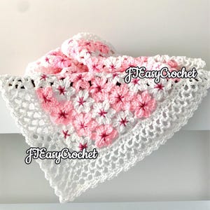 May include: A white and pink crocheted blanket with a floral pattern. The blanket features a white border with a delicate openwork design. The center of the blanket is adorned with pink and white flowers. The text "JEasyCrochet" is visible on the blanket.