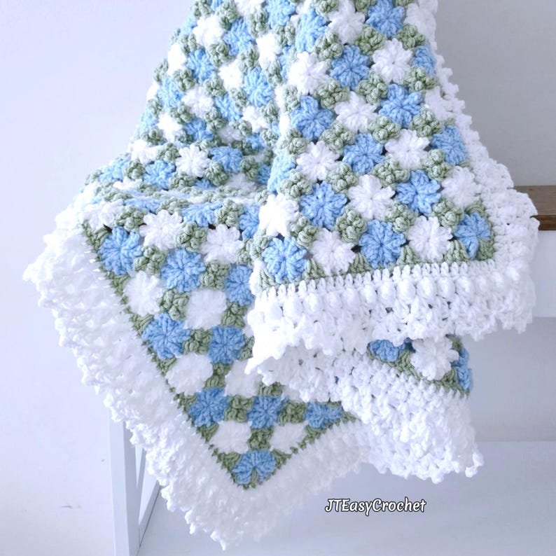 Beautiful blue and white crochet floral blanket made of cornflower motifs is hanging from a display. The blanket resembles a field of white and blue cornflowers framed with a beautiful lace edging.  The lettering says "JTEasyCrochet" meaning that JTEasyCrochet has the best crochet patterns for flower blankets.