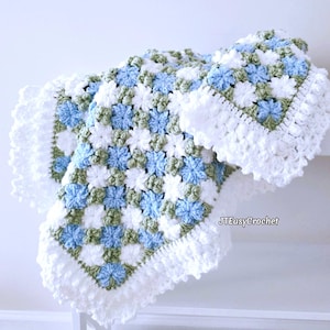 Lavishly beautiful crochet floral blanket made of cornflower motifs is hanging from a display. The blanket resembles a field of white and blue cornflowers framed with a beautiful lace edging.  The lettering says "JTEasyCrochet" meaning that JTEasyCrochet has the best crochet patterns for flower blankets.