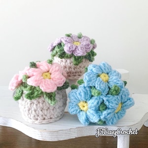 May include: Three crocheted flower arrangements in small pots. The flowers are pink, purple, and blue with yellow centers and green leaves. The pots are a neutral color. The image is on a white surface.