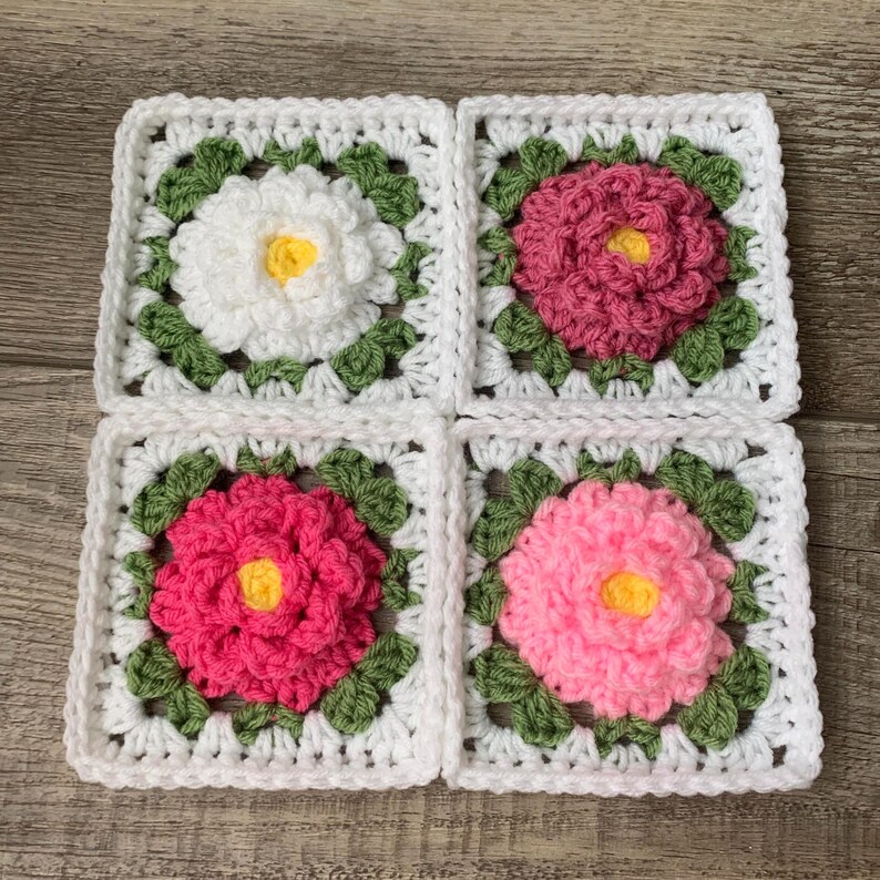 Set of four 3D white granny squares with raised borders laying on a flat surface. Each granny square has a white or shades of pink rose or peony with 2 layers of petals surrounded with green leaves.