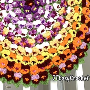 May include: A crocheted blanket featuring a vibrant floral design. The blanket showcases rows of colorful pansies in shades of purple, white, yellow, orange, and burgundy, set against a green background. The text "JTEasyCrochet" is visible.