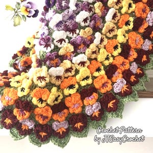 A handmade crochet blanket featuring a vibrant floral design. The blanket showcases a variety of colorful pansies in shades of yellow, orange, purple, and white, with green leaves. The text "Crochet Pattern by JTEasyCrochet" is visible.