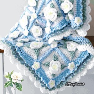 Beautiful crochet granny square blanket is hanging from a display on a light grey background. Each white granny square has a white textured gardenia flower in the center, framed with pale green leaves and light frosty blue border, so the whole blanket remind of a frozen gardenia blanket. There are also bunches of gardenia flowers in the image to highlight the likeness. The lettering logo says: " JTEasyCrochet"